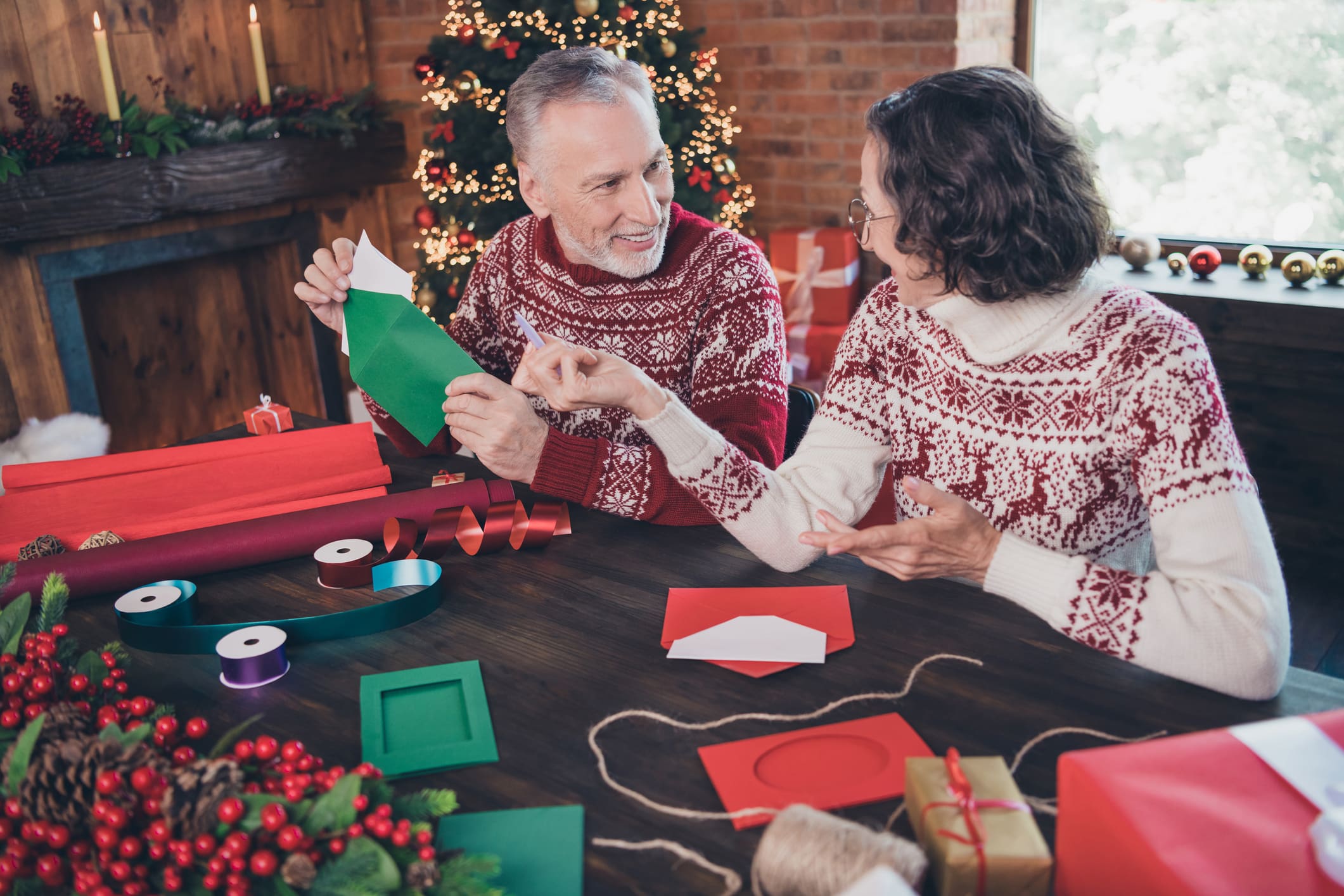 Photo of happy positive couple talking, grandma and grandpa while packing envelop letter wearing ornament sweaters home indoors